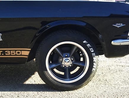 Close-up of a black classic car's rear wheel with prominent white lettering on the tire and a 'GT. 350' marking above it.