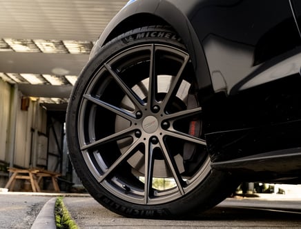 Close-up of a car's back wheel featuring a TSW rim and a Michelin tire, parked in a garage with sunlight filtering through a metallic roof creating shadows.