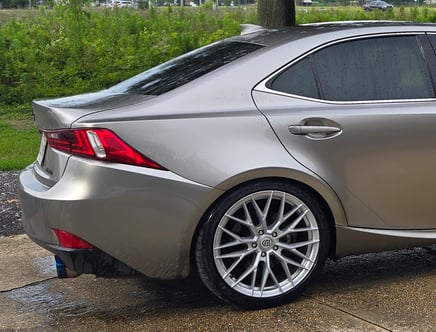 Rear side view of a silver car parked on a wet driveway. The car features shiny, multi-spoke alloy wheels, a red brake light, and tinted windows. Green foliage is visible in the background.