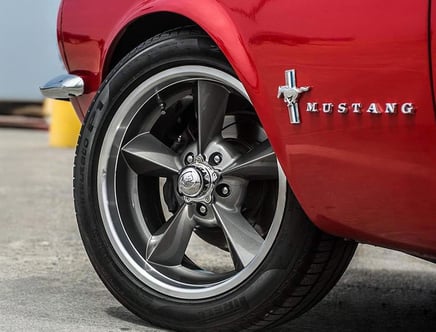 Close-up of a red Ford Mustang's front wheel and emblem, featuring a polished alloy rim and a visible Mustang logo on the car's side panel.