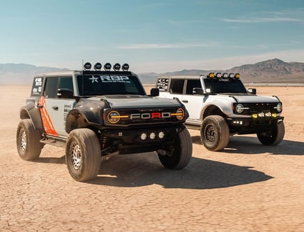 Two off-road trucks with "FORD" on the grille driving in a desert with rugged terrains in the background.