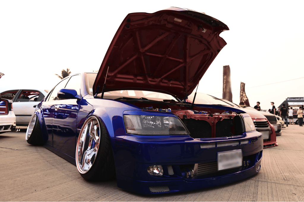A blue customized car with a lowered body and an open hood, showcasing its engine components and stance fitment parked at an outdoor car event.
