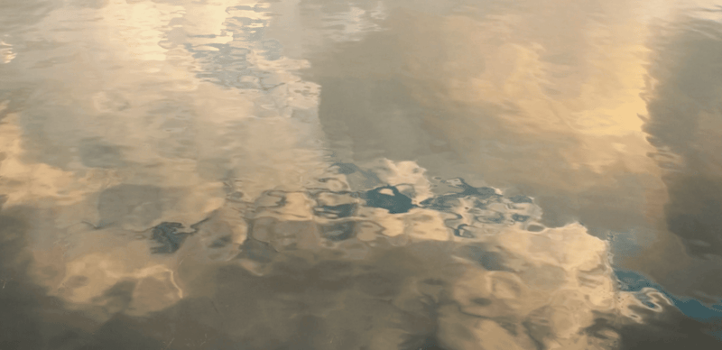Réflexion déformée du ciel et des nuages dans l'eau — teintes dorées et bleues ondulent à la surface.