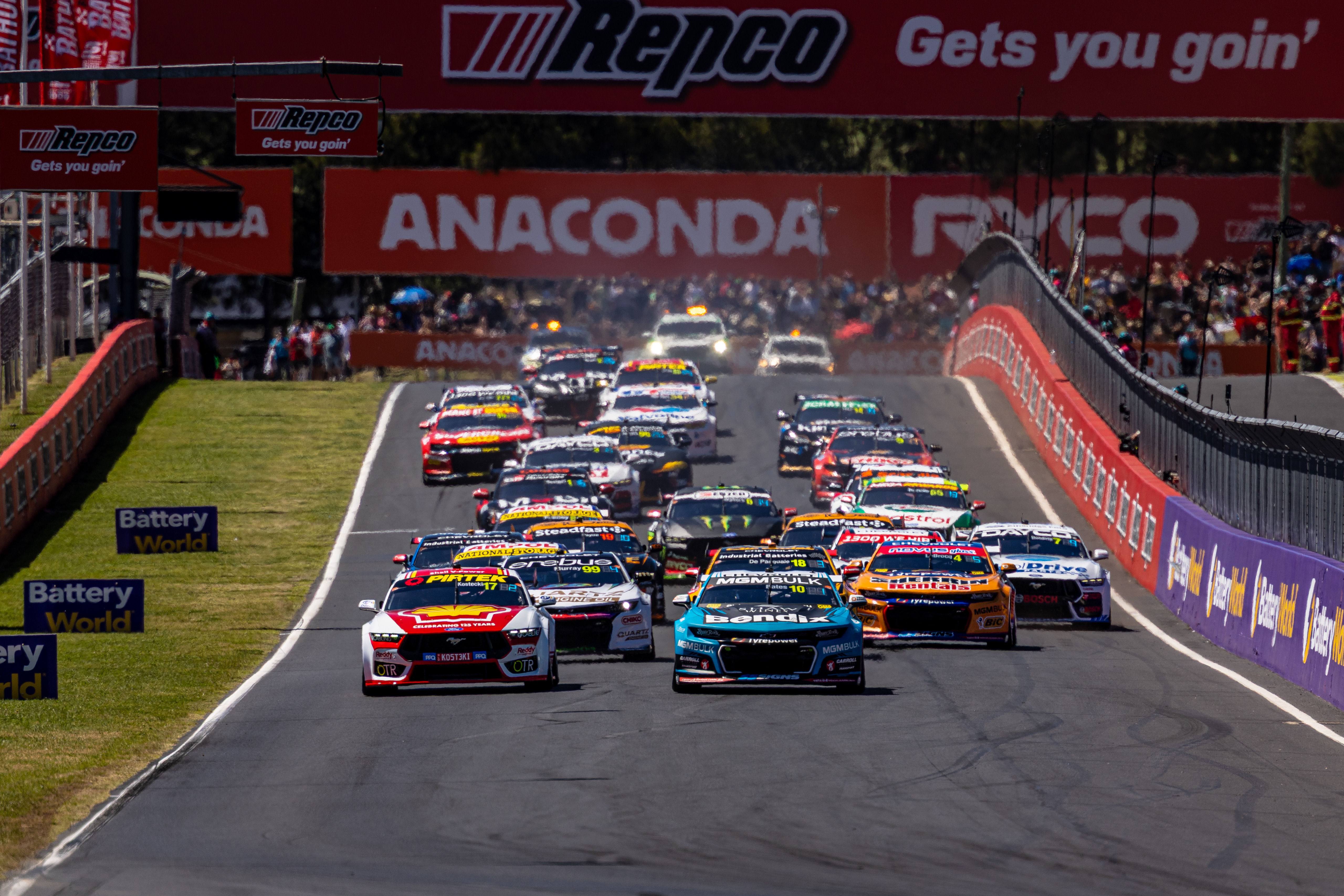 Supercars racing pack descends Mount Panorama led by a white Ford Mustang at Bathurst.