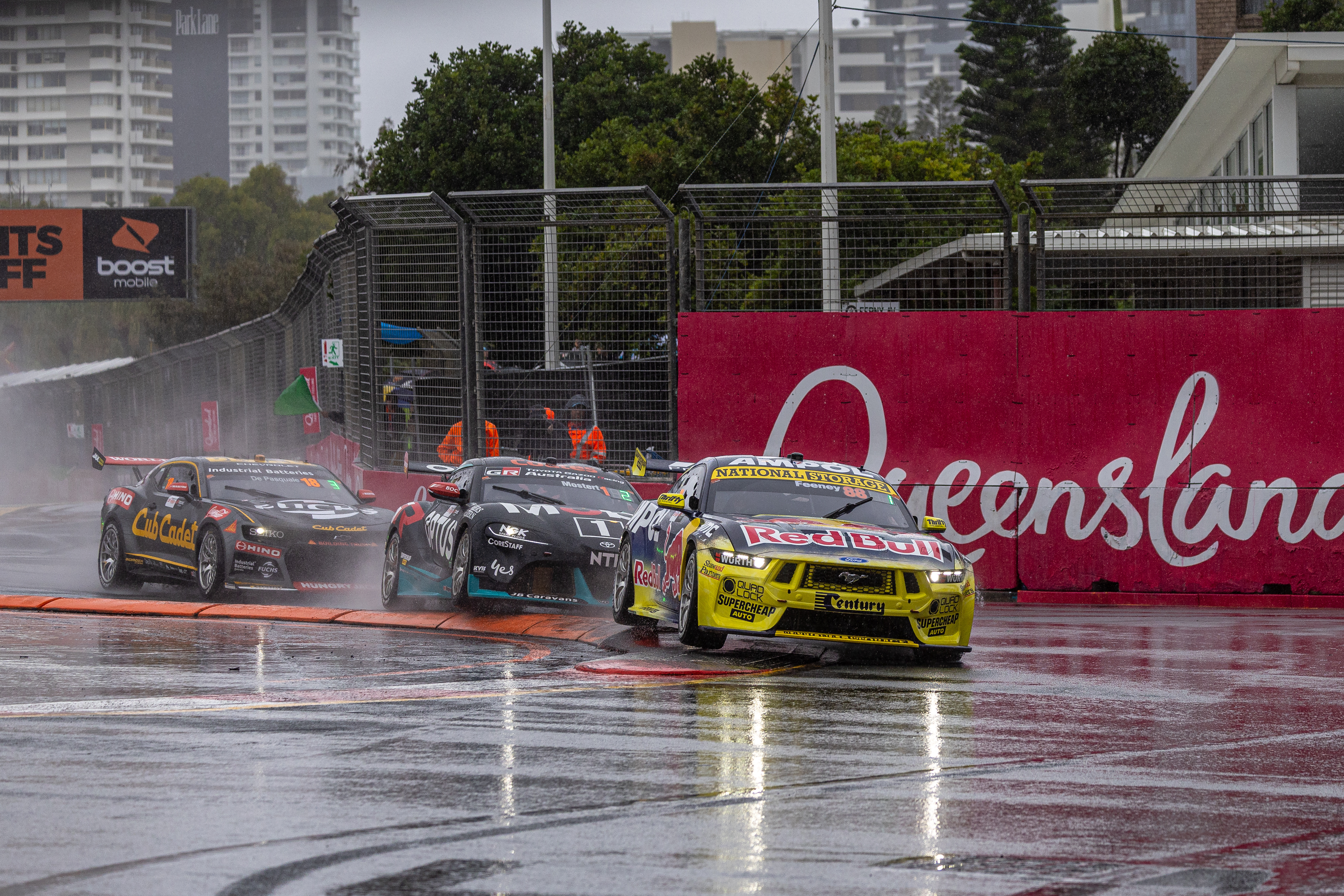Three Supercars round a wet hairpin corner, led by a yellow Red Bull liveried Mustang.