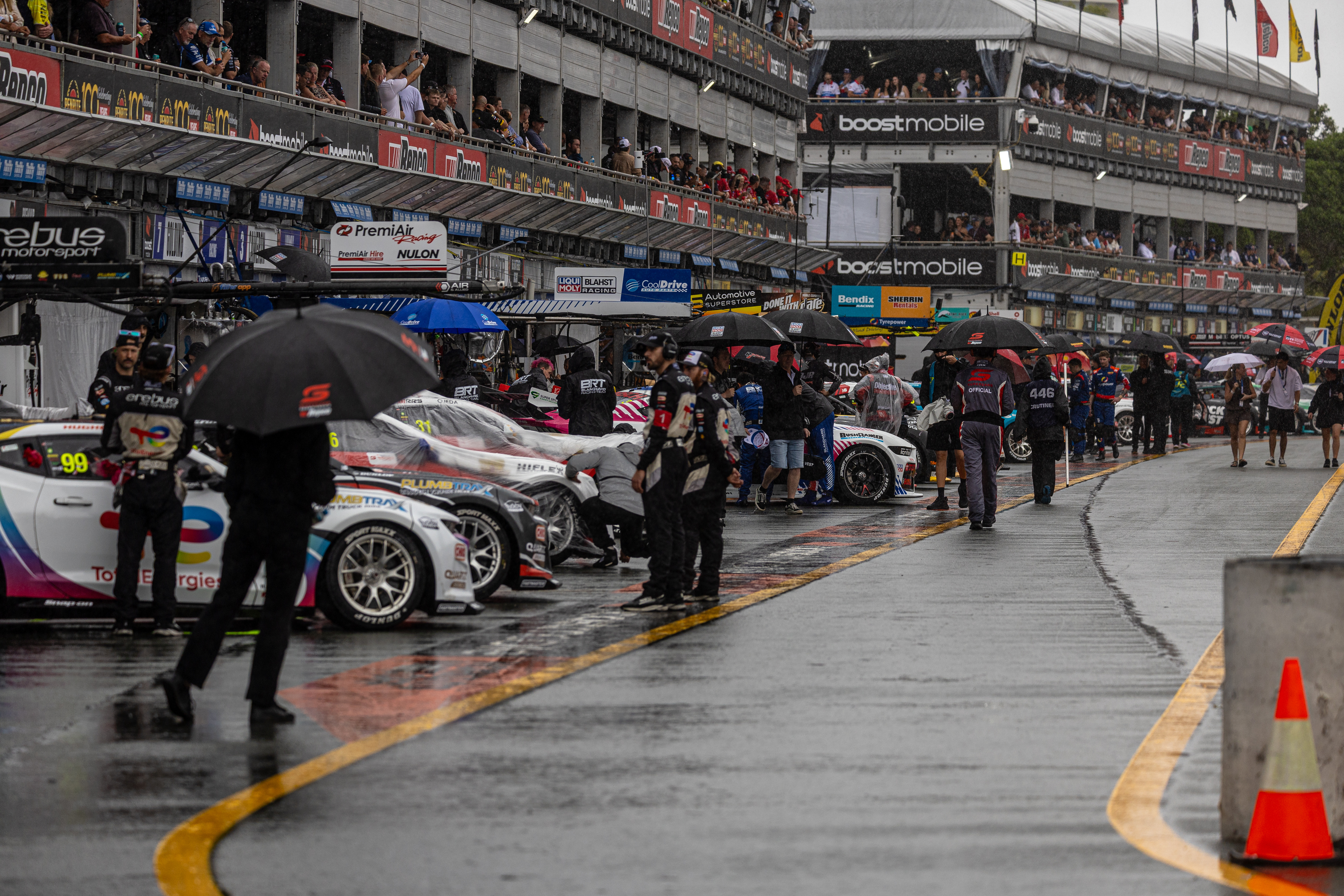Rain-soaked pit lane with race cars, crew holding umbrellas, and crowded grandstands above.
