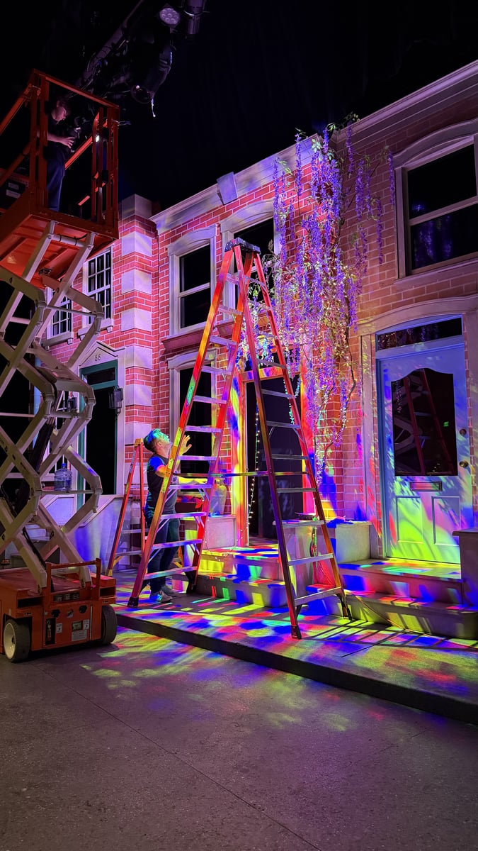 A focus call in progress, a crew member up an A-frame ladder beside a scissor lift, with multicolored gobo breakup scattered across the brick-street set.