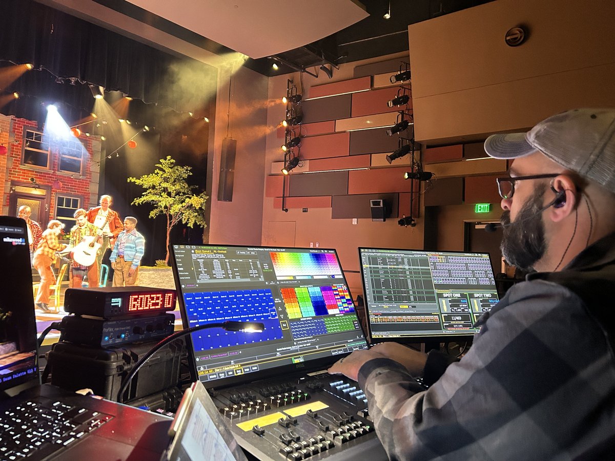 Mike Wood programs the show at an ETC Ion XE console during tech as performers rehearse on the warmly lit street set behind him.