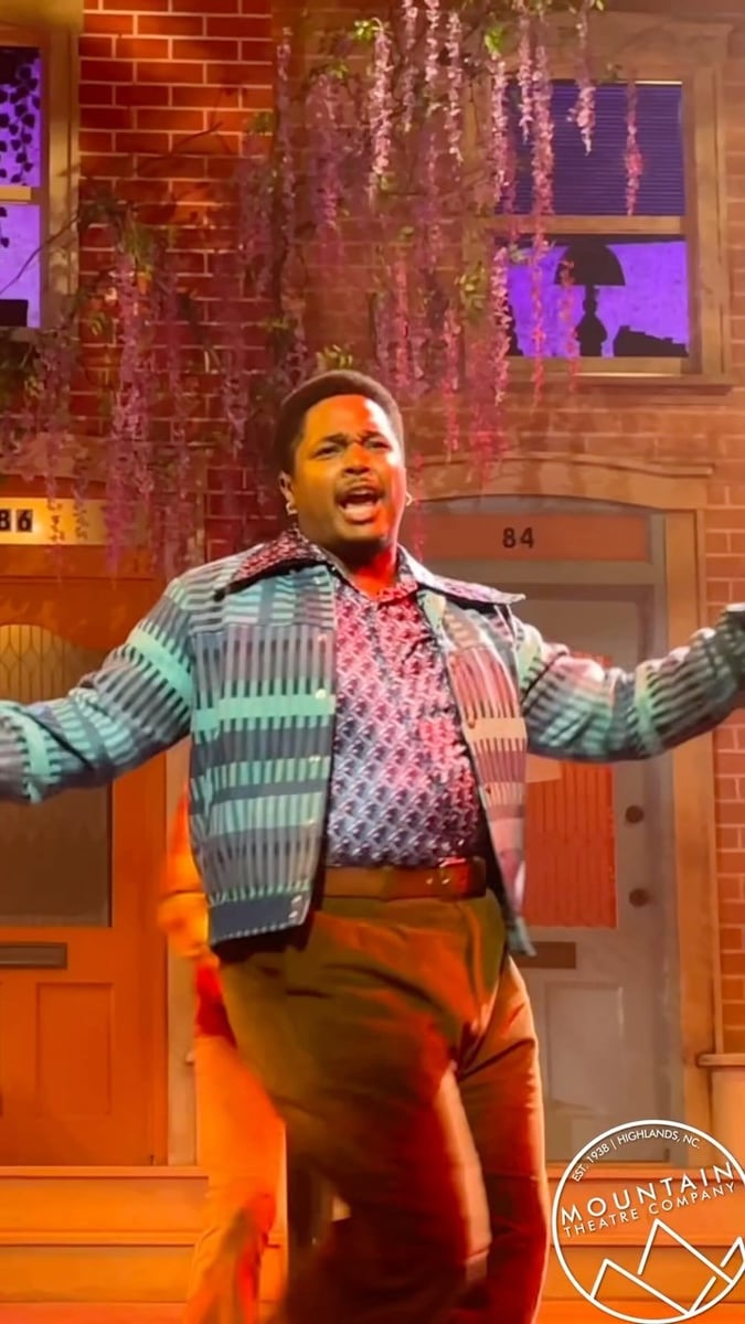 Dancin' In The Street: a performer sings downstage on the street set in a patterned cardigan.