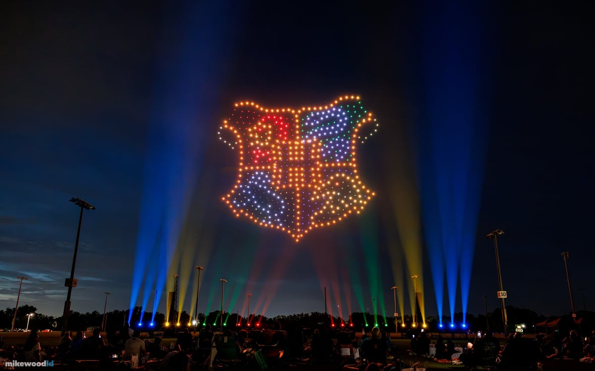 The four-house crest rendered in red, green, blue, and amber drones with blue, gold, green, and red beams rising from the field perimeter.