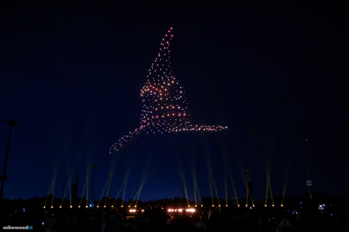 A wizard's hat drone formation in warm rose tones against the deep blue sky, gold beams low along the field.