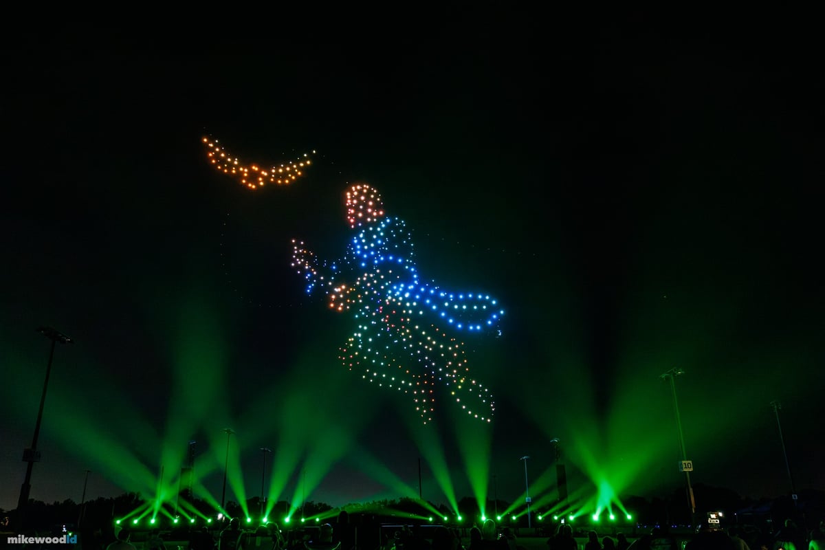 A broomstick rider in blue and amber drones stretching toward a streak of gold, green beams sweeping the field below.