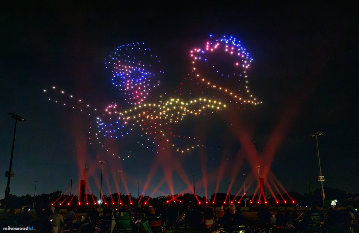 Drone-sketched figures in blue, pink, and gold above the field, red beams crossing in an X pattern below.