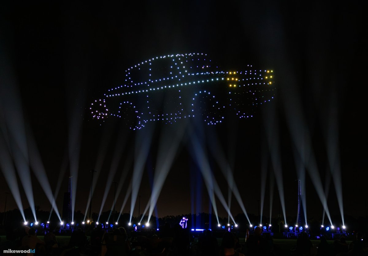 A flying car outlined in white and blue drones with gold headlights, white beams fanning up across the field.