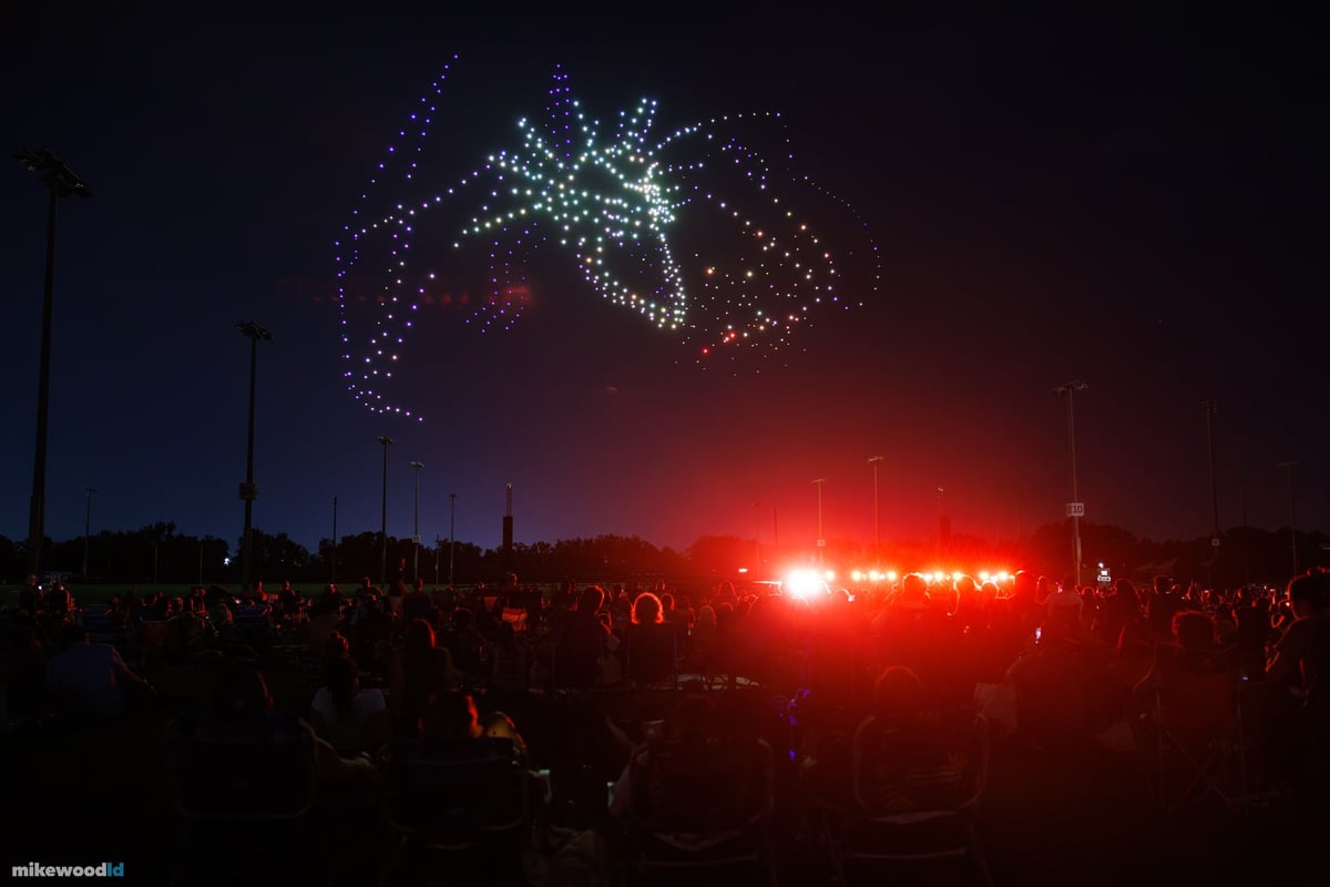 A leaping stag in silver-white drones trailing violet swirls over the audience, red fixtures glowing at the field's edge.