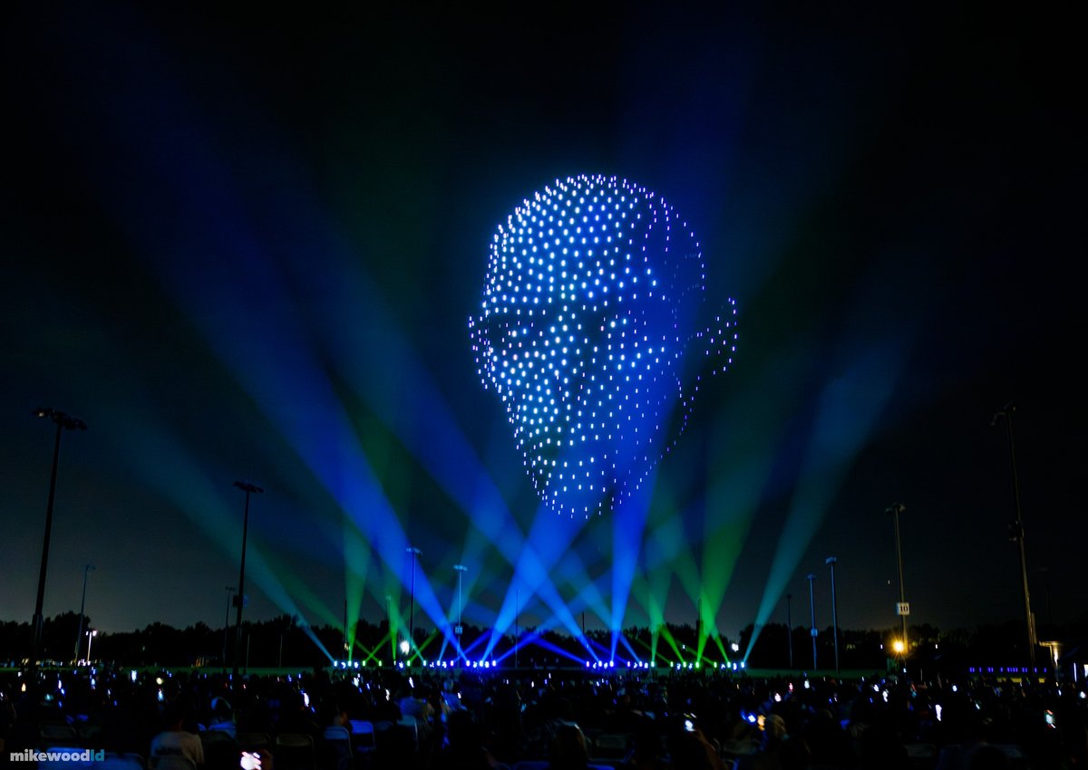 A towering character portrait in ice-blue drones, blue and green beams fanning up from the field.
