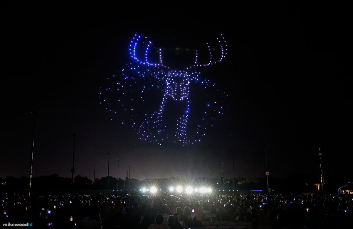 A stag with full antlers rendered head-on in blue and violet drones, white fixtures blazing at the center of the field.