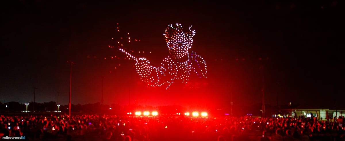A drone-rendered figure with arm outstretched trailing amber embers, the field and audience washed deep red.