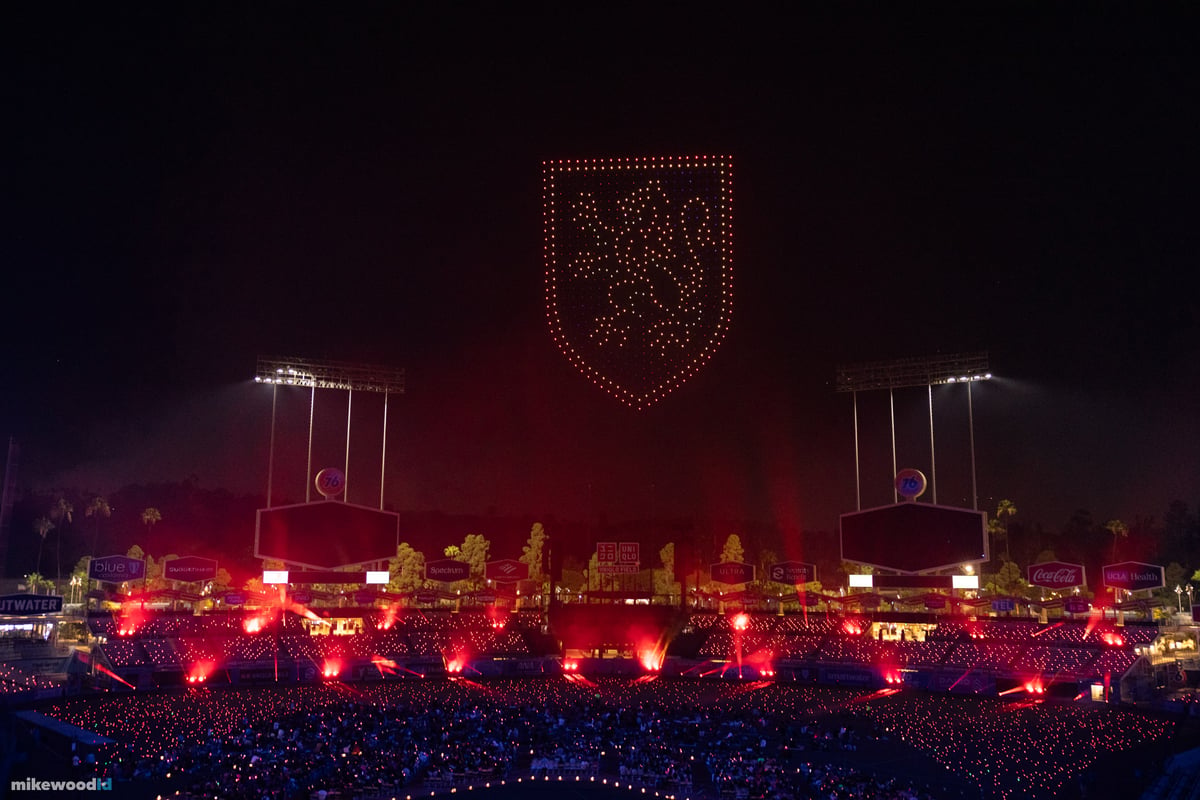 A house crest in red and amber drones above the field as red pyrotechnic flares erupt along the stage front, the bowl washed red.
