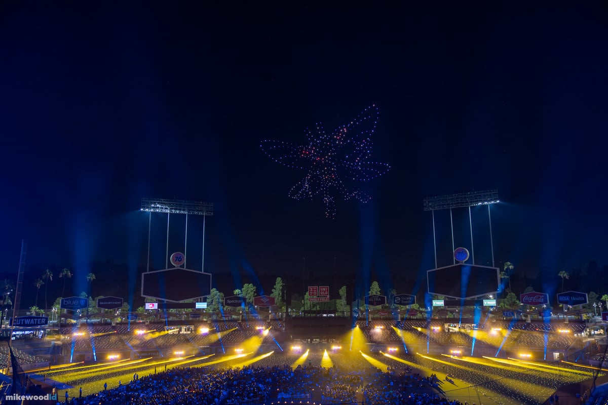 A bright starburst formation in blue and red drones above the stage, blue and amber beams fanning out in radial lines below.