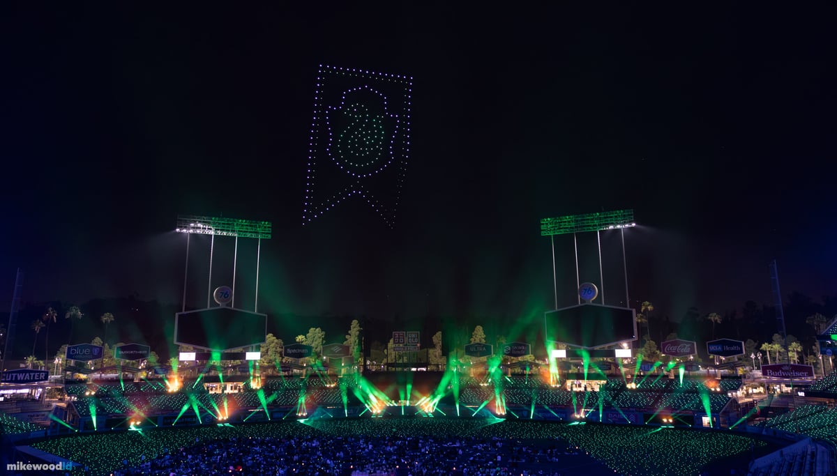 A green house banner rendered in drones over the stadium, green beams fanning up from the stage and crowd.