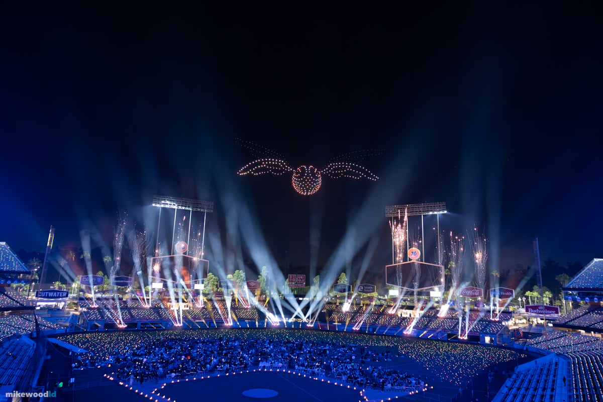 The Golden Snitch rendered in amber drones, seen from high in the stands above a haze-filled Dodger Stadium ringed with white beams.