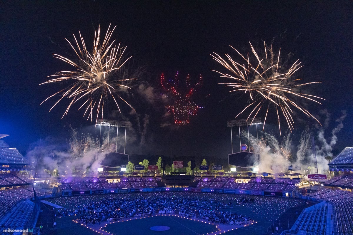 A creature rendered in red drones flanked by two large gold firework bursts above Dodger Stadium, the bowl lit blue below.