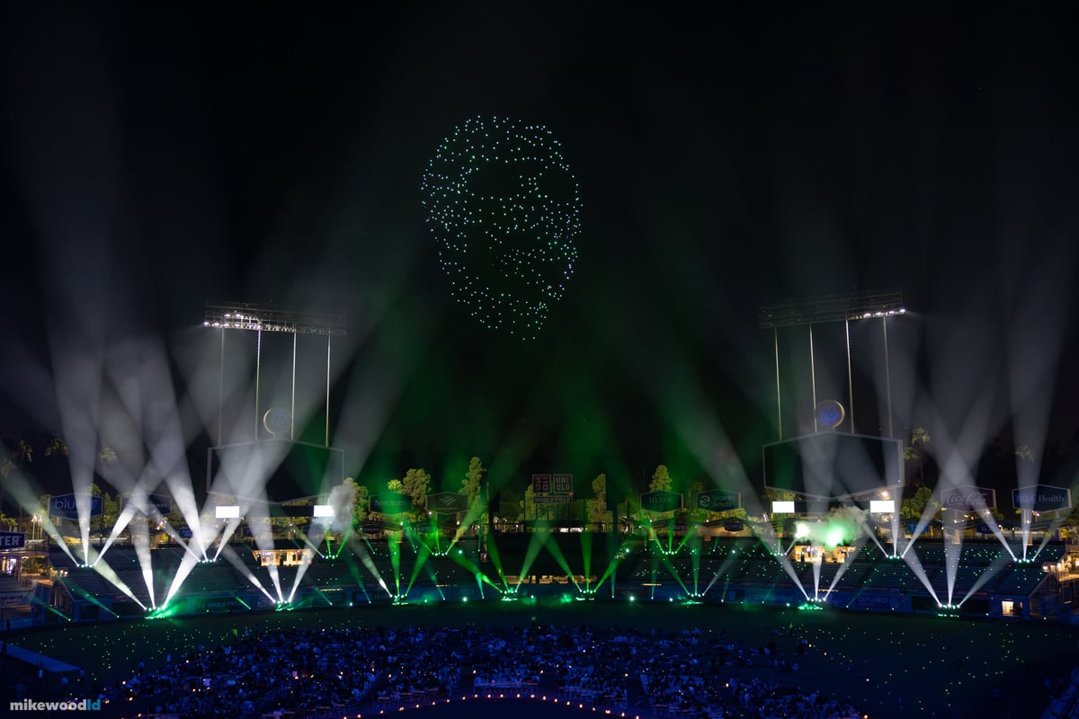 A glowing skull formation in green and white drones above the field, white beams fanning up from the stage in a tight symmetrical spread.