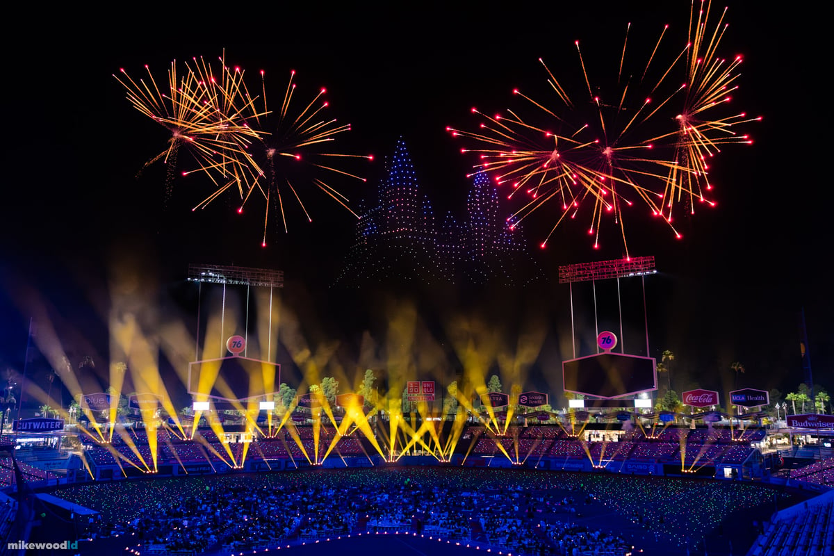 A drone formation framed by gold fireworks and rising sparks above the field, white beams fanning up from a blue-lit crowd.