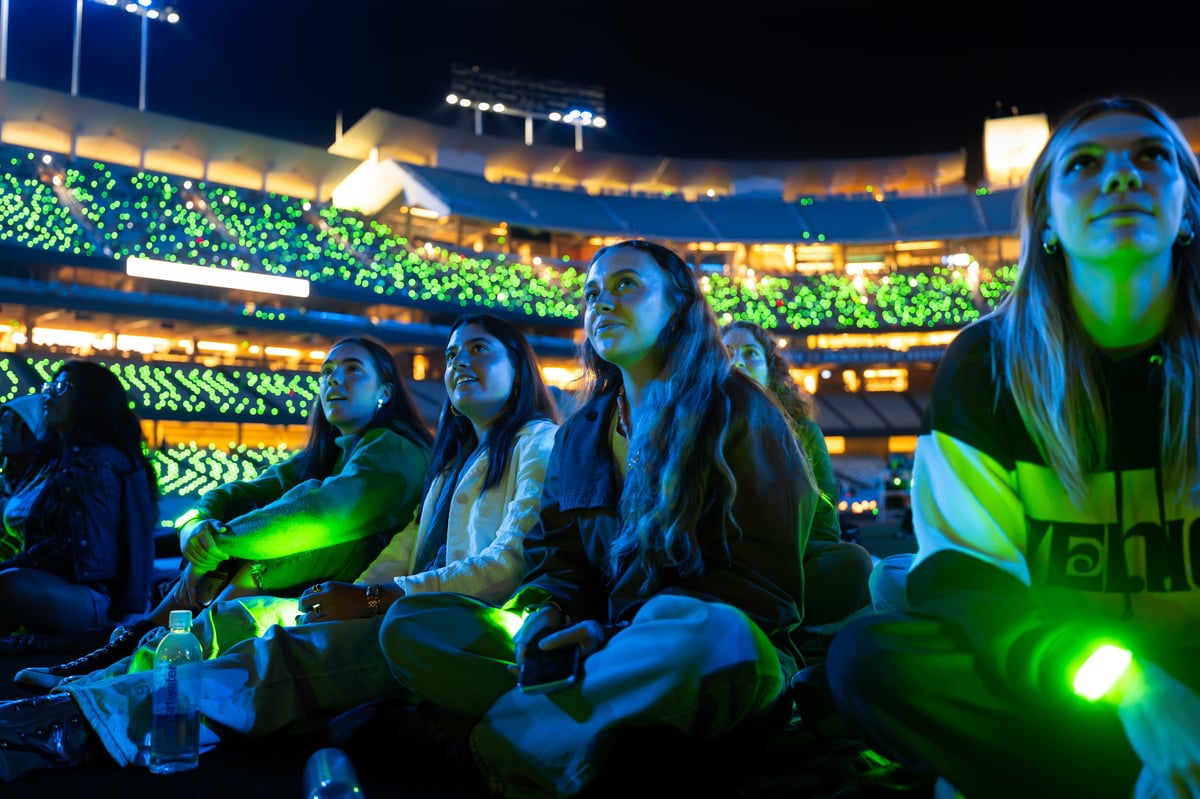 Spectators seated on the field look up at the show, the packed Dodger Stadium stands behind them washed in matching green.