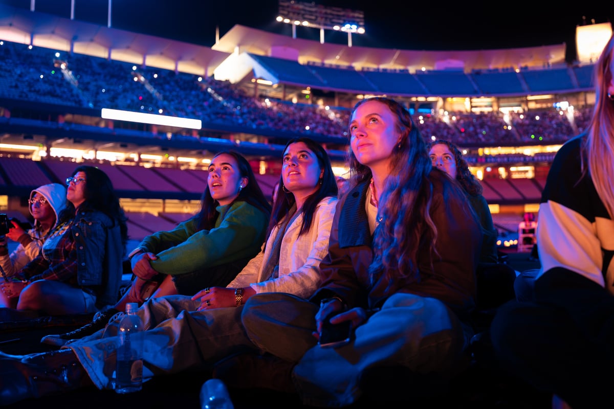 A row of spectators sits cross-legged on the field, their faces and the full stadium bowl lit in blue and pink.