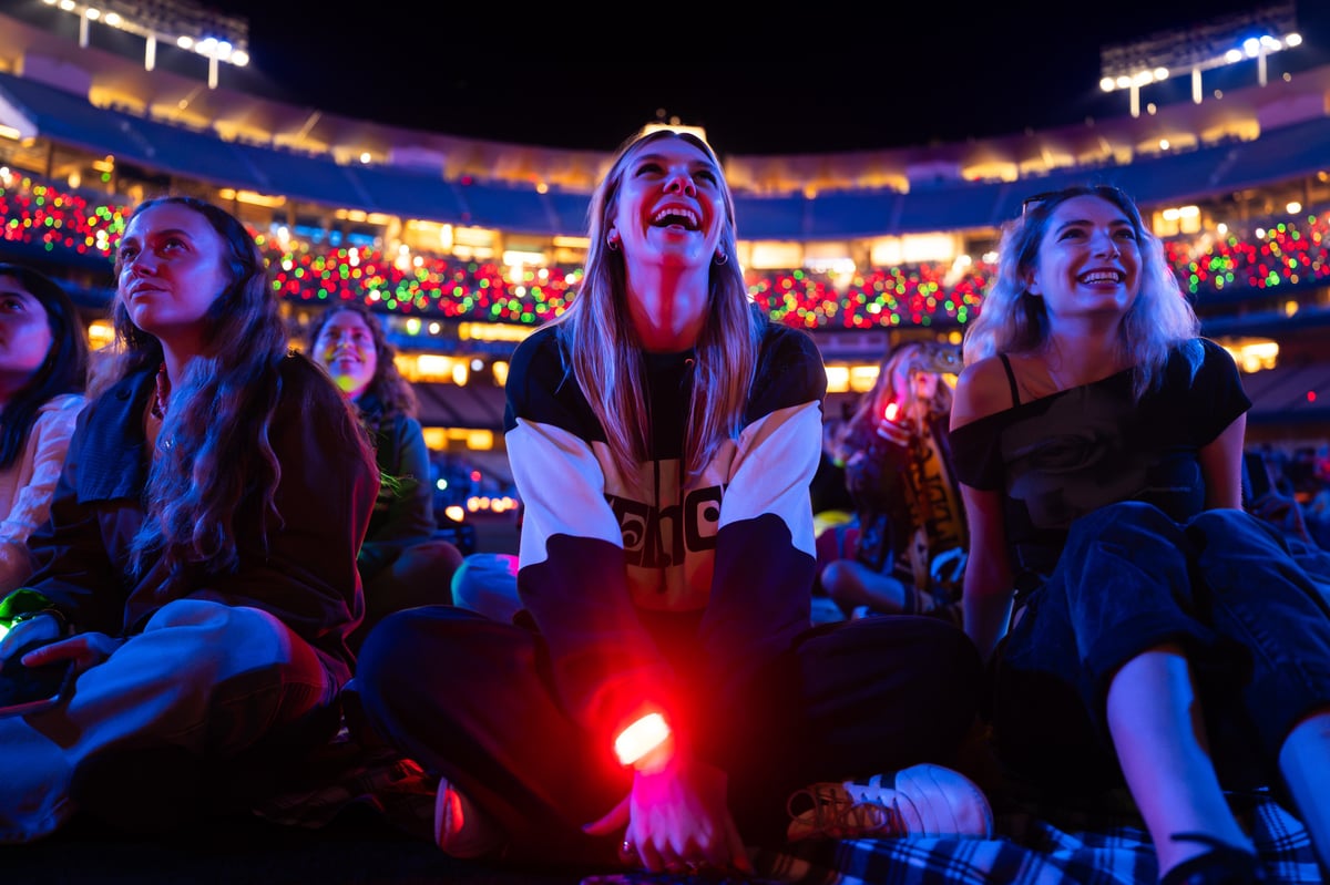 Spectators laugh on the field with red LED wristbands glowing, the Dodger Stadium stands behind them lit in red and green.