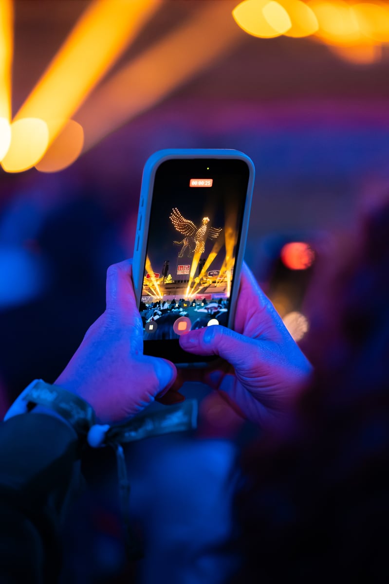 A spectator films the show on a phone, the screen catching a golden phoenix drone formation above the stage.