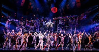 The full company of CATS with arms raised beneath a red moon as Old Deuteronomy presides from the junkyard set