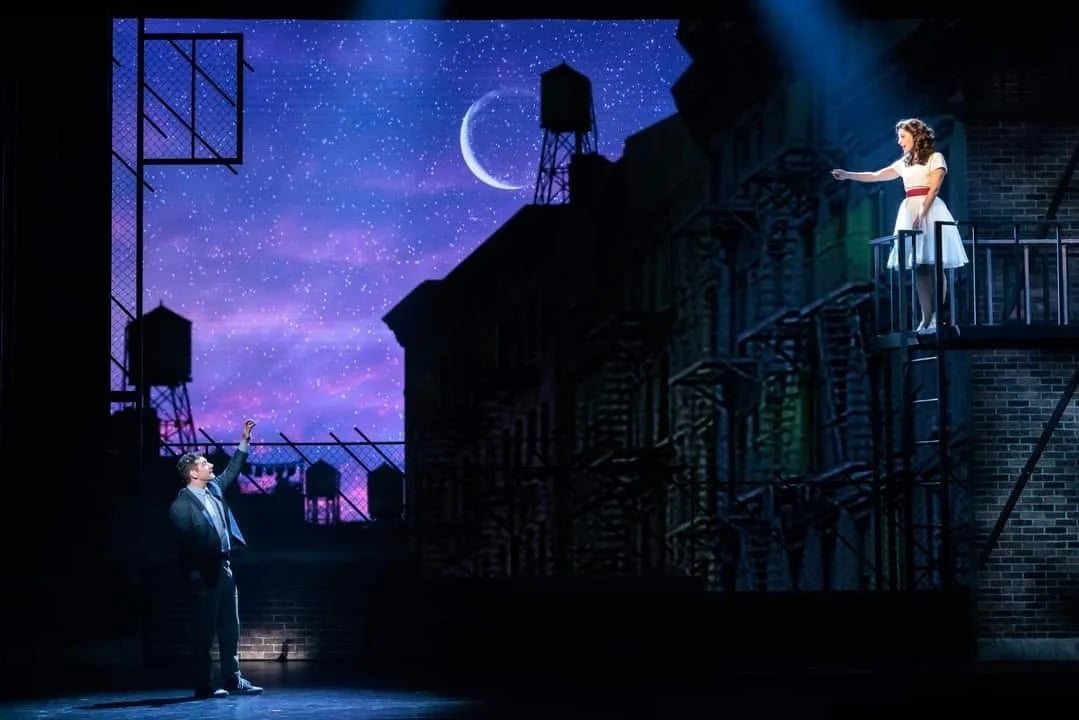 Tony reaches up toward Maria on the fire escape beneath a crescent moon in the balcony scene