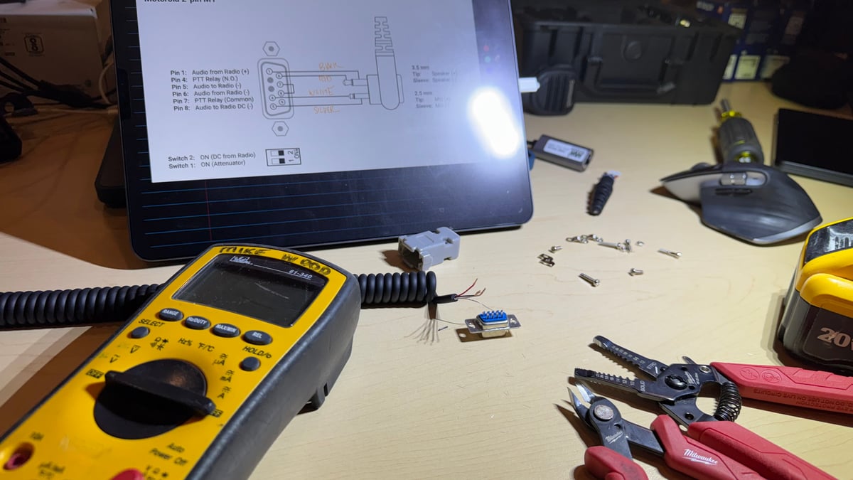A workbench during prep: a tablet showing a connector pinout, a Fluke multimeter, wire strippers, and a comms cable being wired by hand.