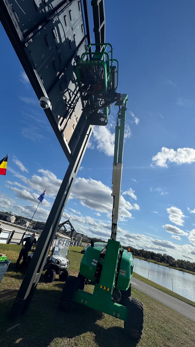 A camera hanging from the underside of a large LED video wall, with an articulating boom lift raised alongside it and the pond behind.