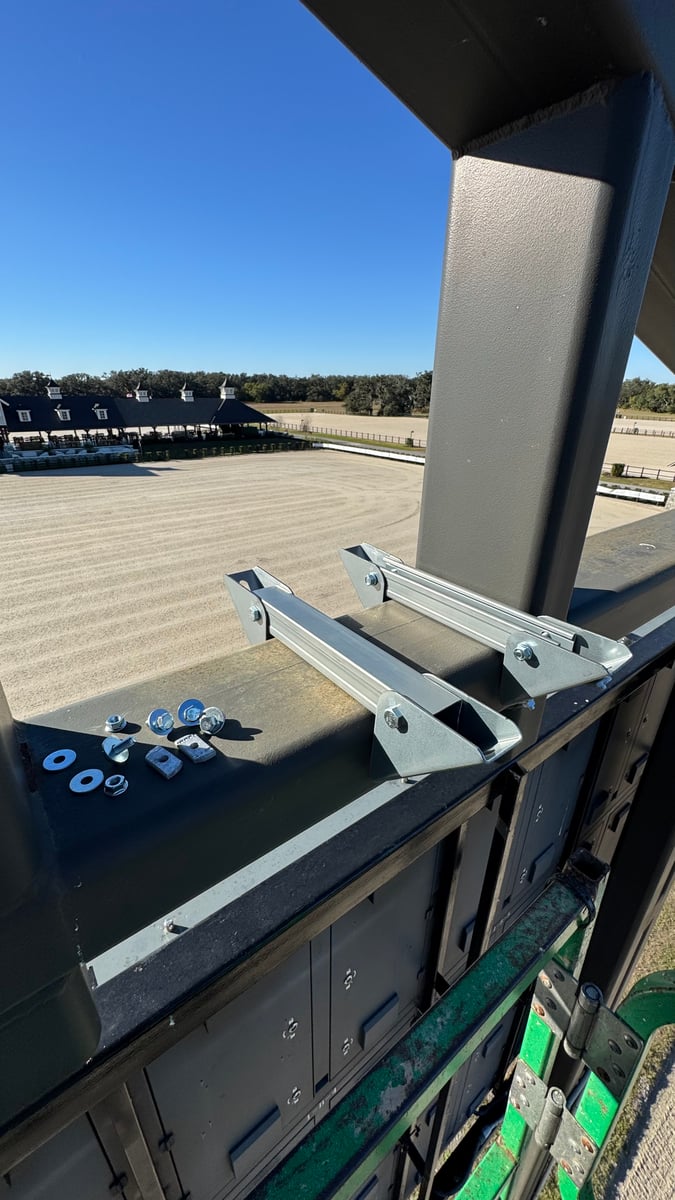 A galvanized steel custom bracket clamped to the railing above a dressage arena, mounting hardware laid out beside it.