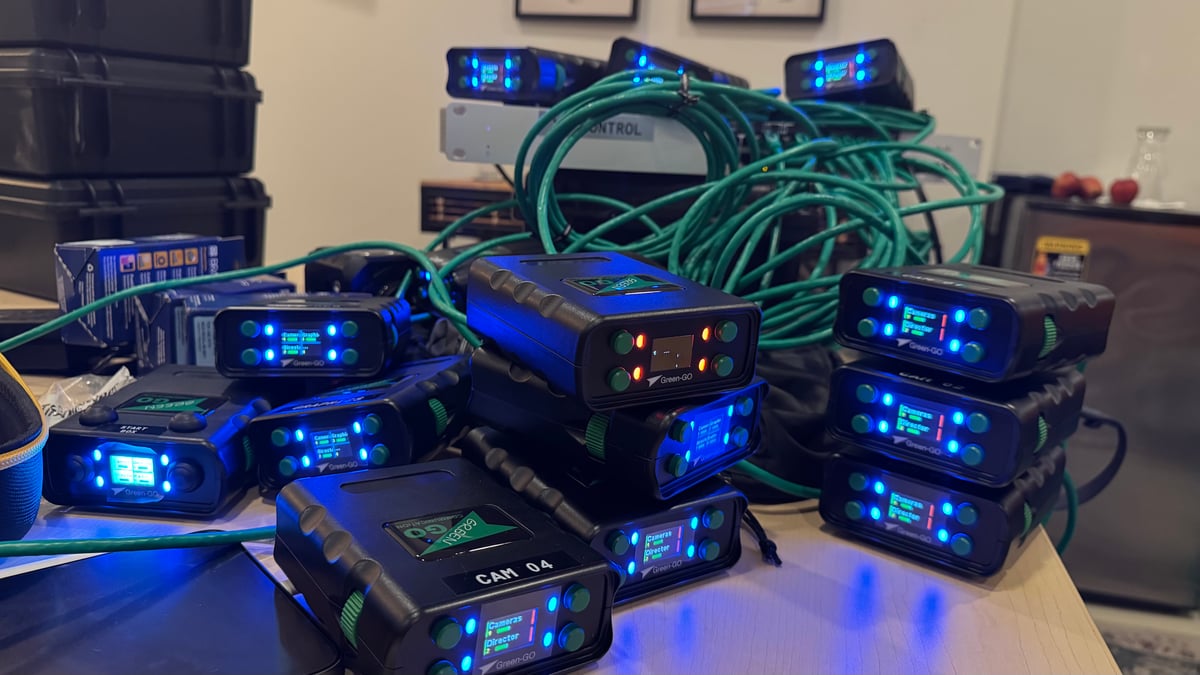 A pile of Green-GO intercom beltpacks with lit blue screens and coiled green Ethernet cables on a bench, labeled by camera position.