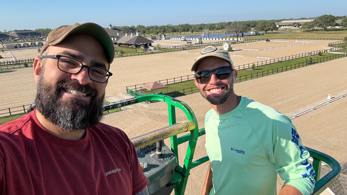 Two men smiling in a raised boom-lift basket, the sand dressage arenas and barns of the equestrian center spread out behind them.