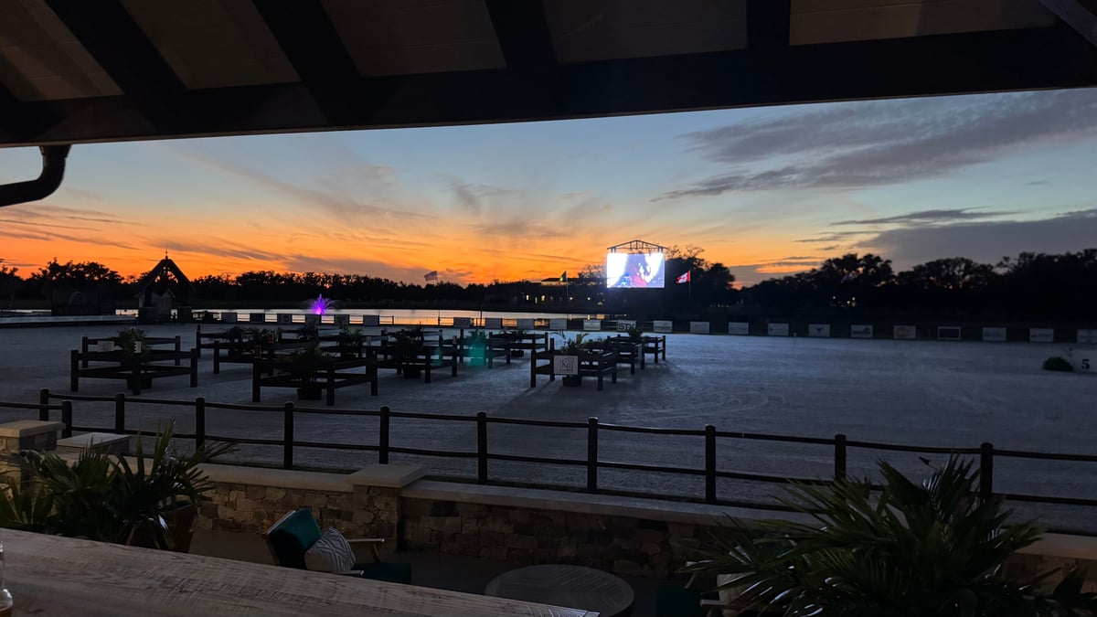A dressage ring at dusk seen from under a shaded pavilion, an orange sky over the tree line and a video screen glowing across the arena.