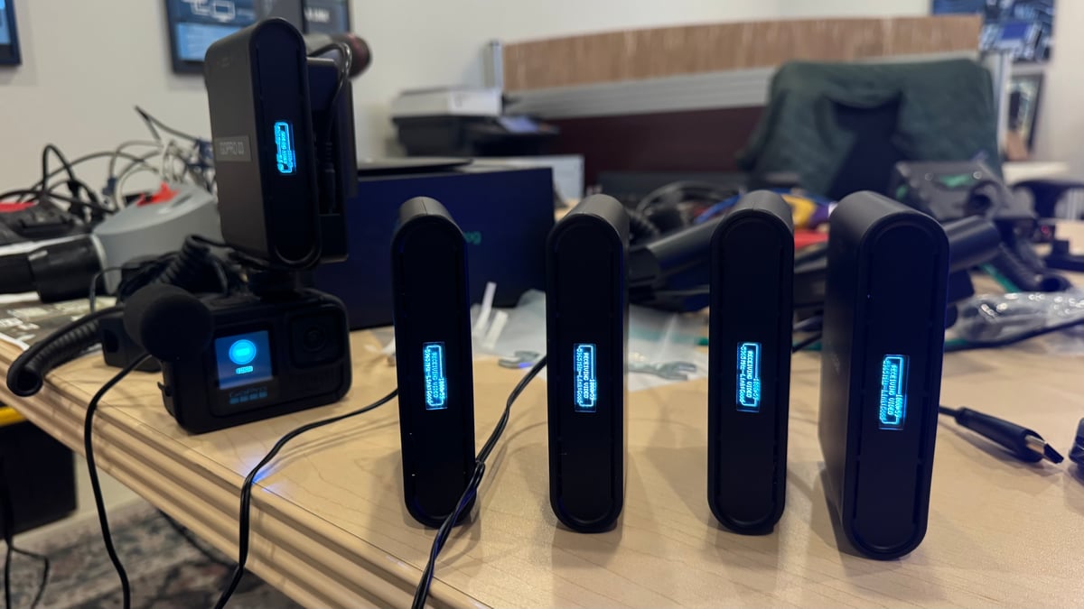 Four small Teradek wireless transmitters standing on a bench with their status screens lit blue, beside a GoPro camera on a charger.