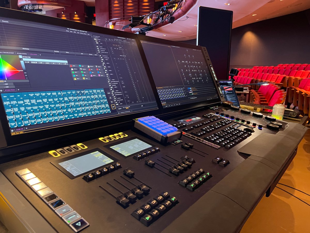 Wide view of the Eos programming desk in the Cobb Energy booth, Eos console with dual displays showing magic sheet and dynamic FX palette, red theater seats visible at left