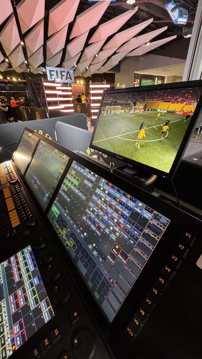 grandMA2 layout touchscreens at the lighting position, with a lit FIFA sign and a live match on the monitor behind.