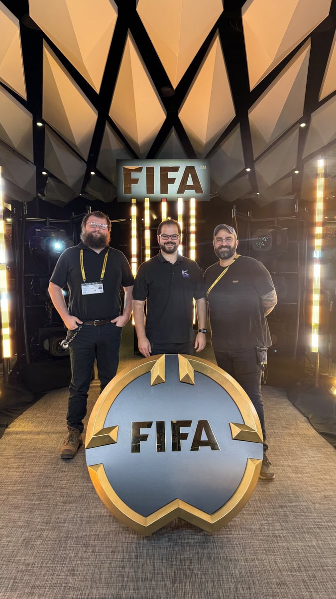 Three crew members at the lighting position, one in a Kinetic Lighting shirt, posed in front of a FIFA crest and sign.