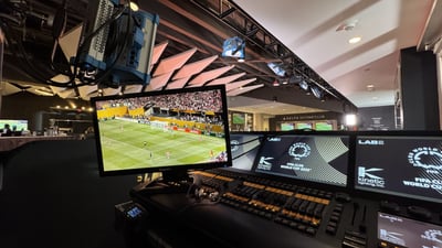 Lighting control position in a stadium suite: a grandMA2 console with Kinetic Lighting layout screens, overhead moving lights on truss, and a monitor showing the live FIFA Club World Cup match.