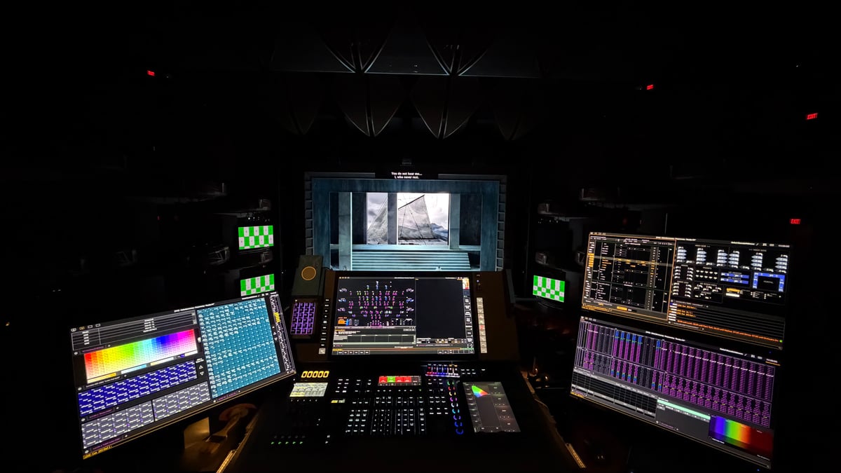 Wide head-on view of the lighting programming booth at Cobb Energy during Götterdämmerung tech, Eos console centered, dual XKeys panels flanking, multiple displays, stage scrim visible upstage