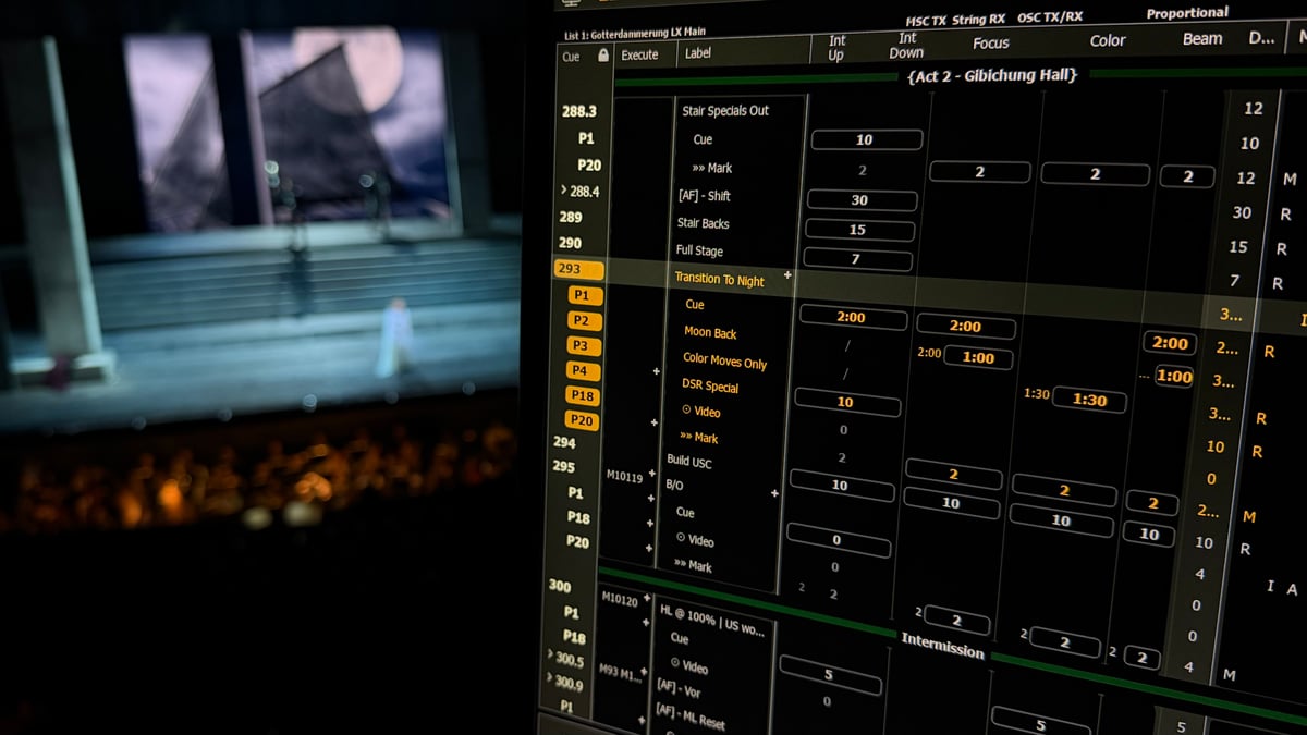Close-up of the Eos cue list display showing Götterdämmerung Act 2 Gibichung Hall cues (Transition To Night, Moon Back, Color Moves Only, DSR Special) with the stage visible blurred at left and a singer on the staircase