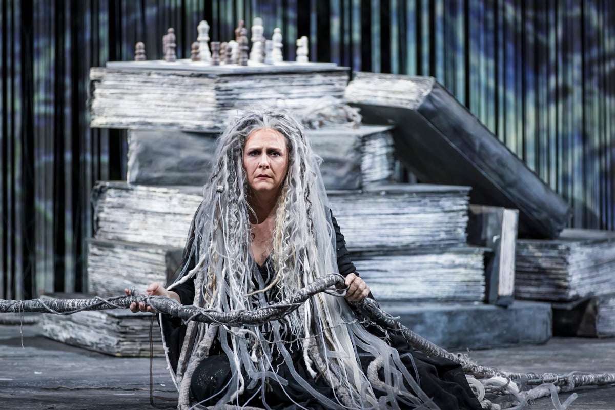 A Norn sits with the rope of fate in her hands, giant weathered books and a chess set stacked behind her.
