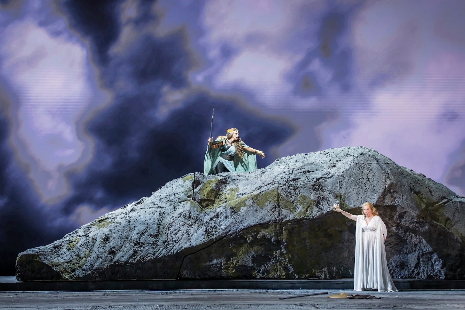 A figure with a spear lunges from the summit of the rock toward a woman in white below, under a full-stage wall of storm-cloud projections.
